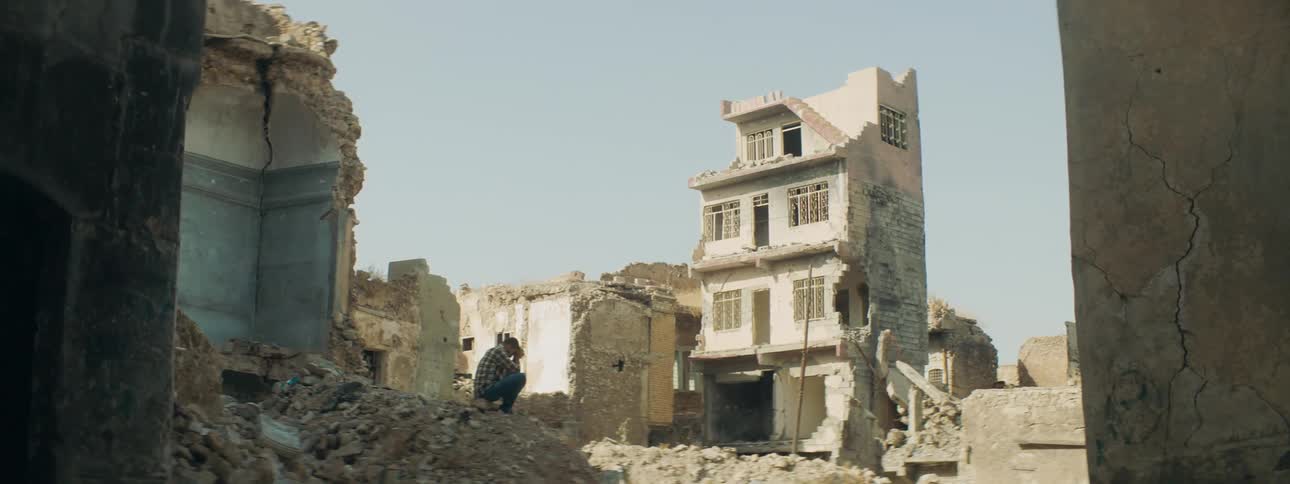 Man sits on rubble with his head in his hands near ruined buildings
