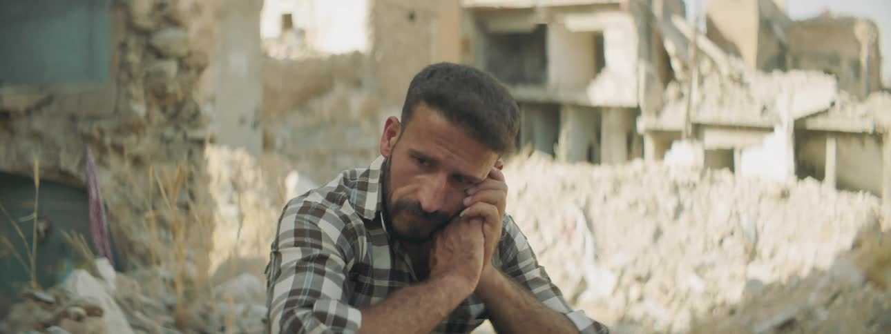 A man sits down amongst some ruble and stares off sadly