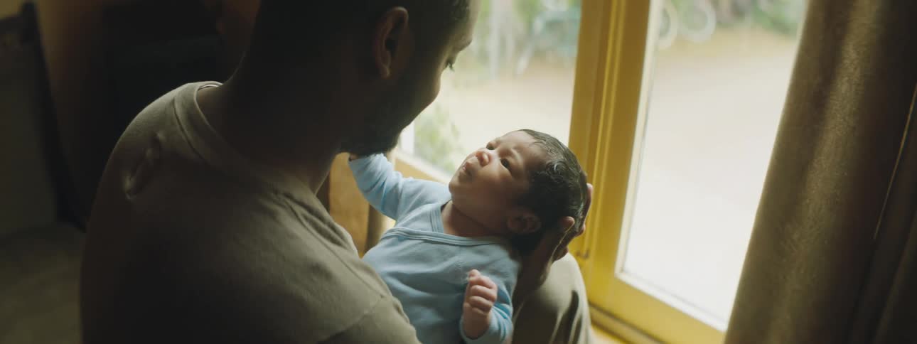 A man sits in front of a window at home and holds his newborn son