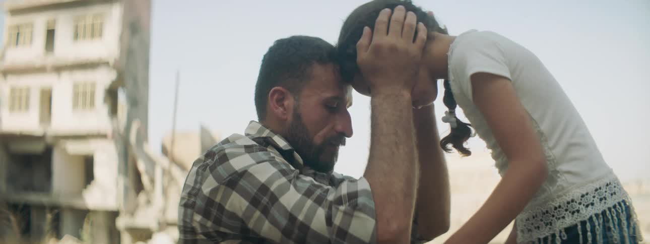 Sitting father kisses and reassures daughter near ruined building and rubble
