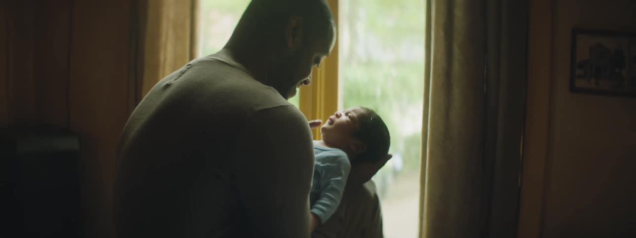 A man cradles a baby and strokes its cheek with his finger in front of a window in a house