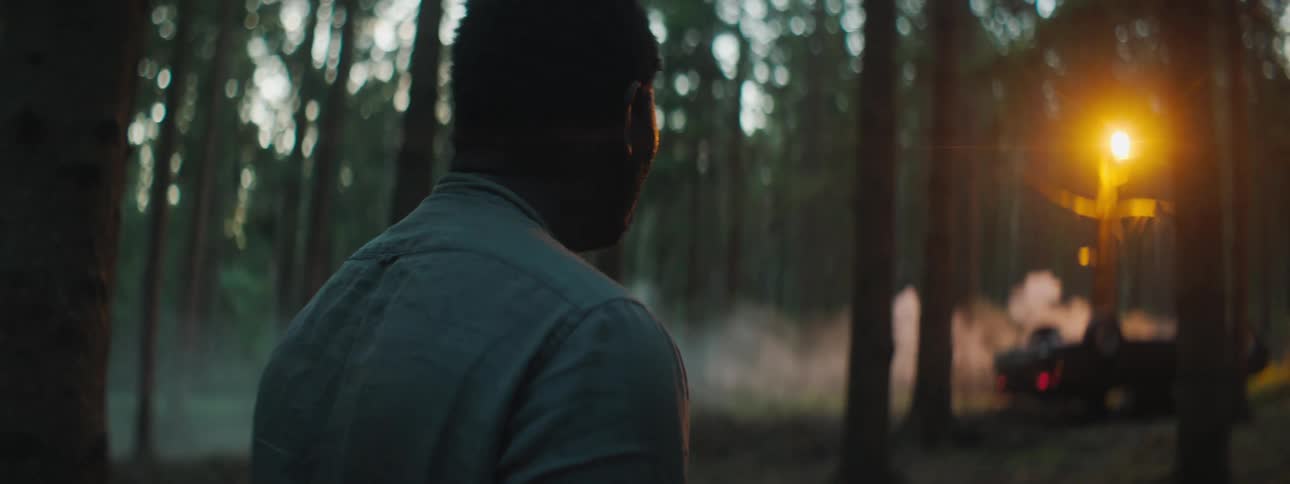 Man stands looking around in forest before walking toward over turned smoking car near streetlight