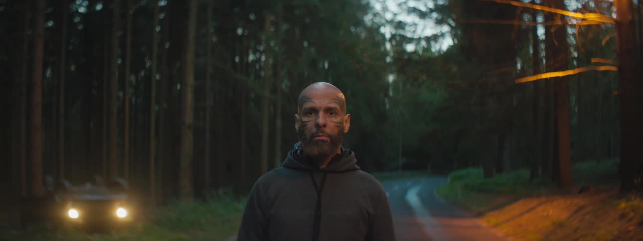 A man standing in the middle of a road next to a forest with his car off to the side
