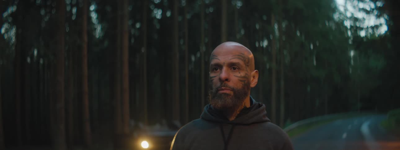 A man with a tattooed face standing on a forest road with a car in the distance