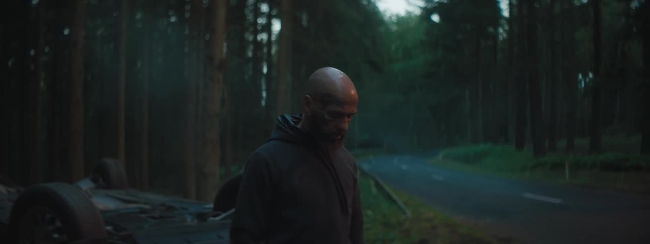A male standing near a flipped over car near a roads and woods