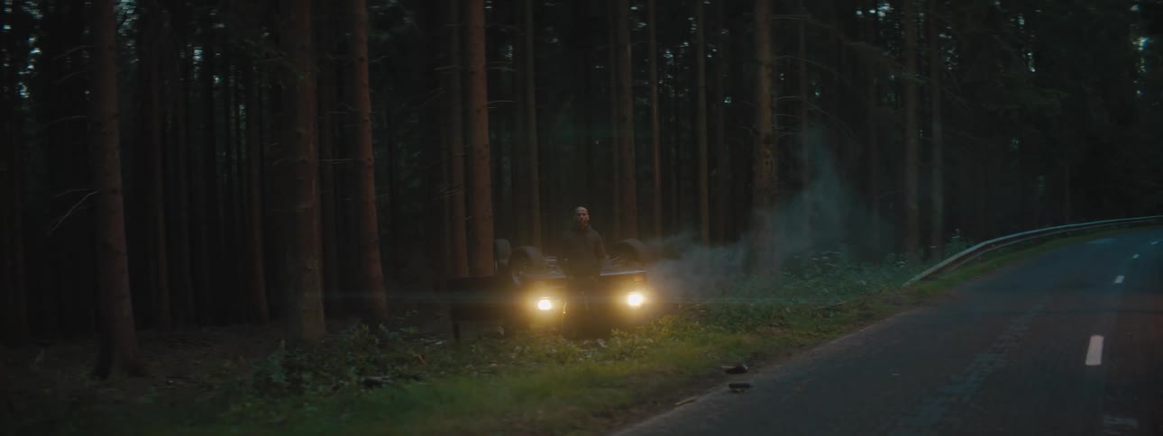 A person standing in front of a flipped over vehicle in the side of the road with trees in the background