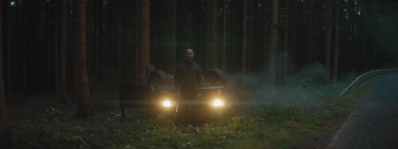 A man standing in front of a flipped over vehicle in the side of the road with trees in the background