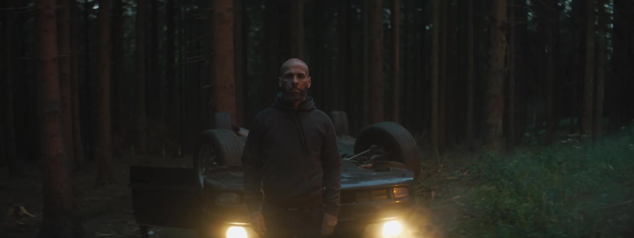 A man standing in front of an upside down car in a forest after an accident