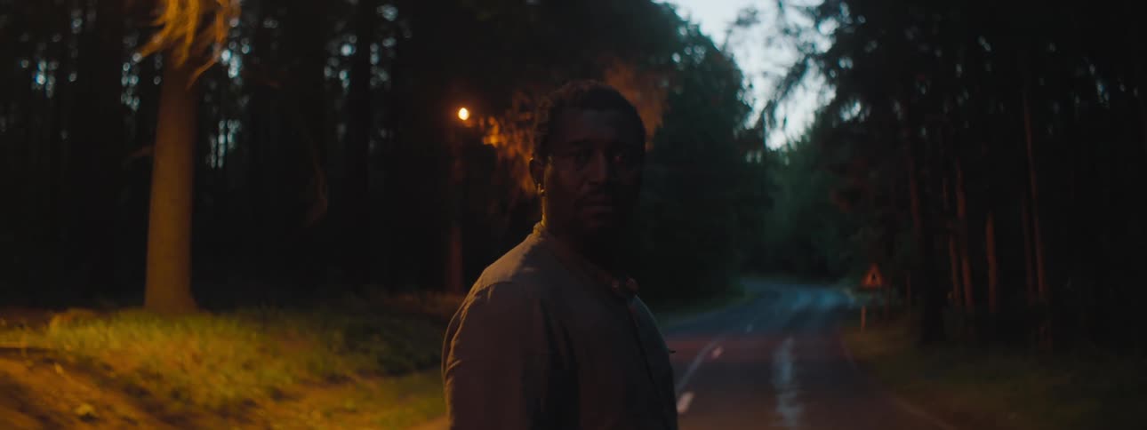 Depressed man with a shirt stands in the middle of the road in the forest and stares at the camera at dusk