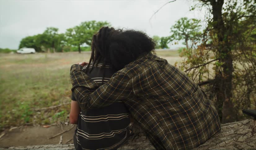 A teenage girl sitting on a tree trunk in a field reaches over and hugs her sister who sits beside her