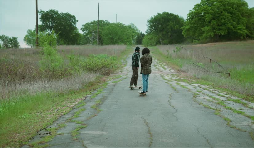 Man with black whooly hat dreadlocks brown pants skateboards on country road with woman with curly hair plaid shirt and jeans