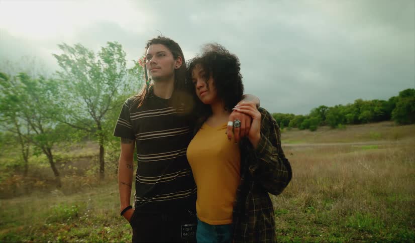 A man and woman embrace while looking into the distance as they stand outdoors on a cloudy day