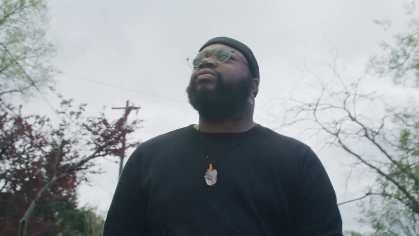 Man with black beanie earring glasses beard and black sweatshirt walks down the street under cloudy sky
