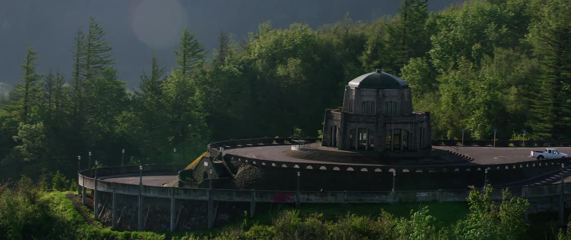 A large building is on a mountain top surrounded by a forest and overlooking a body of water
