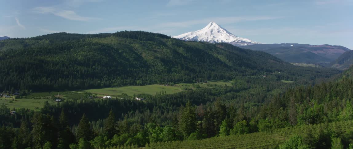 A snow capped mountain peak looms over a vast countryside covered in hills and farmland