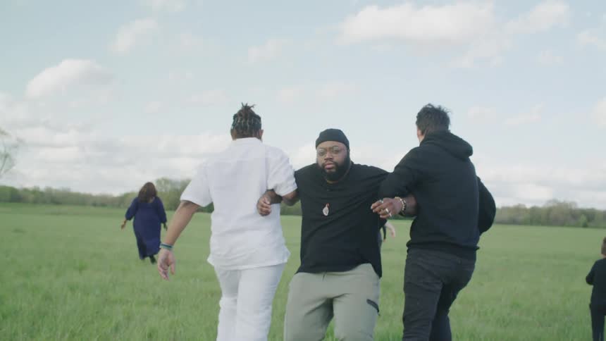People walk and kids skip across a field as another man just stands there staring and two men grab him by the arms and pull him across the field