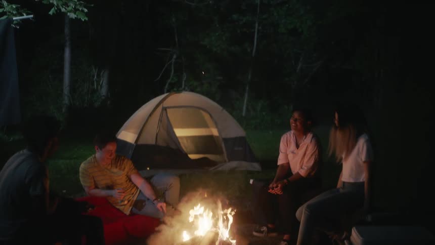 A man sits around a camp fire with another man and two women and pokes a stick in the fire