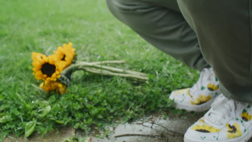 A man is in a park on a pathway and is squatting down crushing sunflowers he is holding