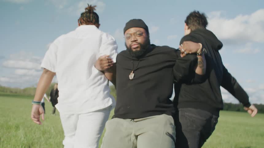A man stands in a grassy field with others walking in it and two men come up and grab his arms and pull him across the field