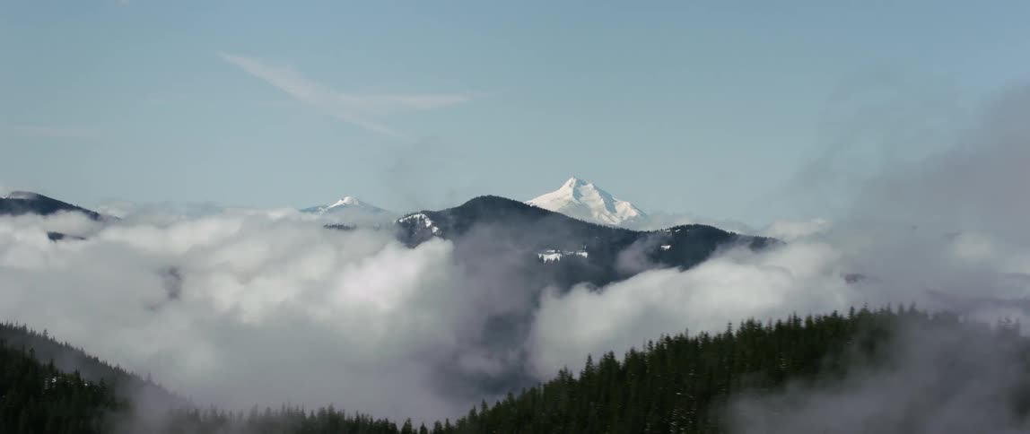 A snow capped mountain peak looms over smaller mountains covered in forest during the day