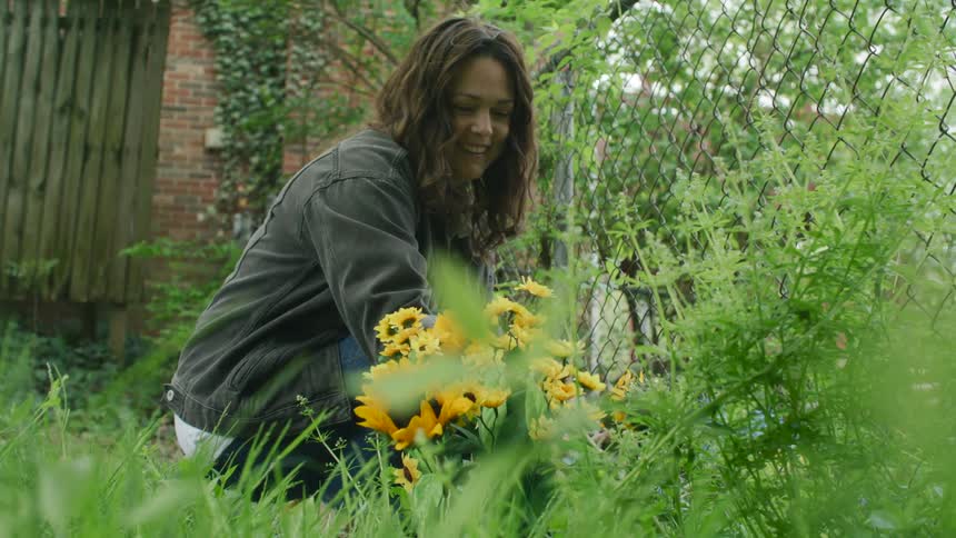 Woman with wavy hair denim jacket and jeans crouching down picks flowers smiling in the garden