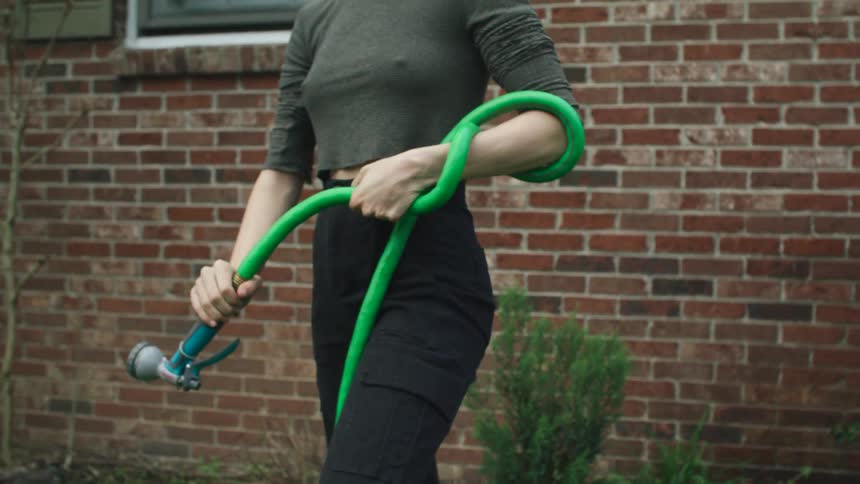 Woman tightly winds a bright green garden hose around her lower arm standing in the backyard of a brick house