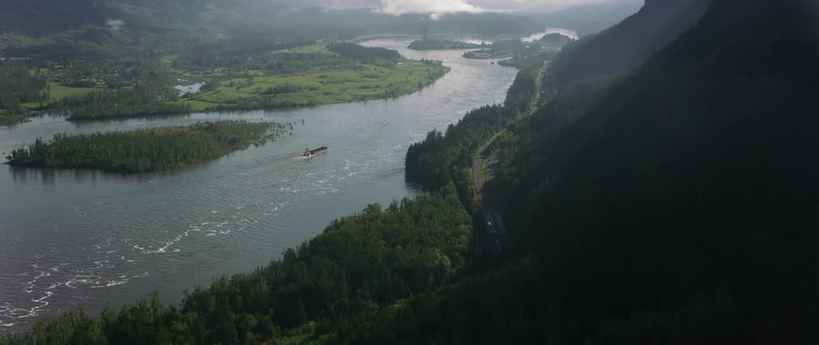 Aerial view of ship traveling on river next to highway and high mountains