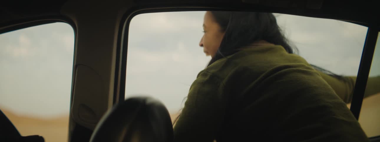 A smiling young woman leans out of the back window of a vehicle as it travels through a desert on a sunny day
