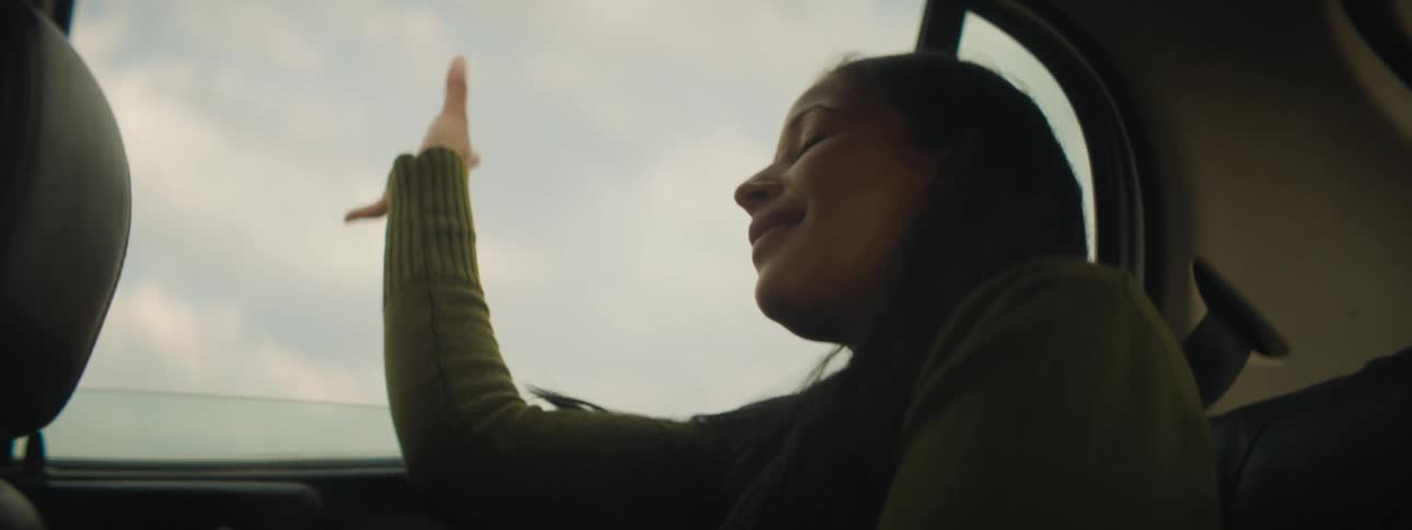 A girl riding in the backseat of a vehicle with the wind blowing on her face with the sky and clouds in the background