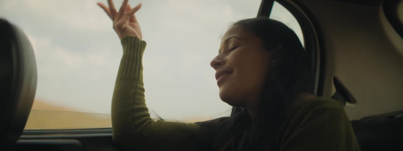 A girl sitting in the back of a vehicle with wind blowing on her face with the sky and clouds in the background