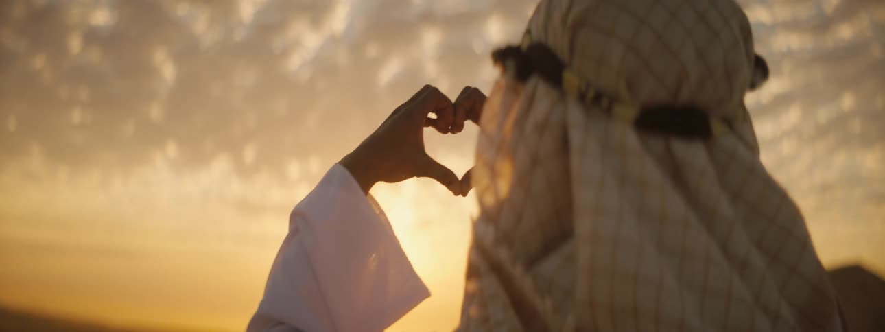 Man wearing a keffiyeh stands over a dune in the desert making a heart sign with his fingers at sunset