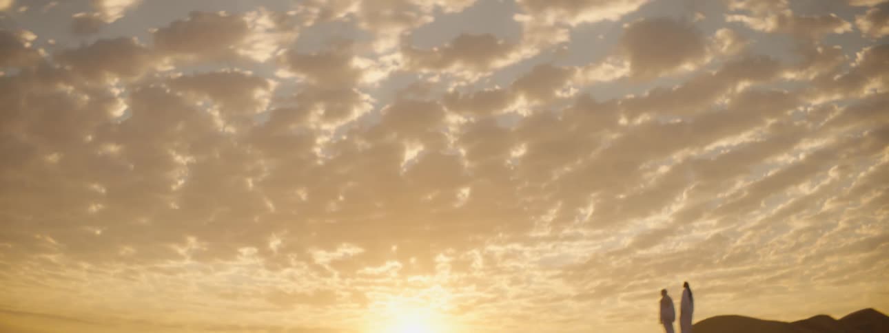 Two people stand on top of a sand dune in a desert as the sun rises