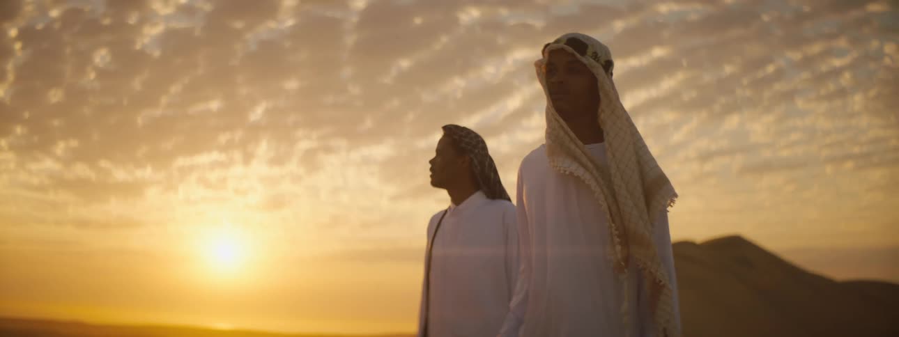 Two men standing in a desert with the sunset sky and clouds in the background