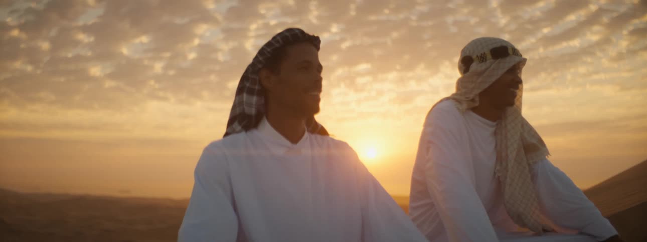 Two men sitting in the desert in robes and turbans as the sun sets