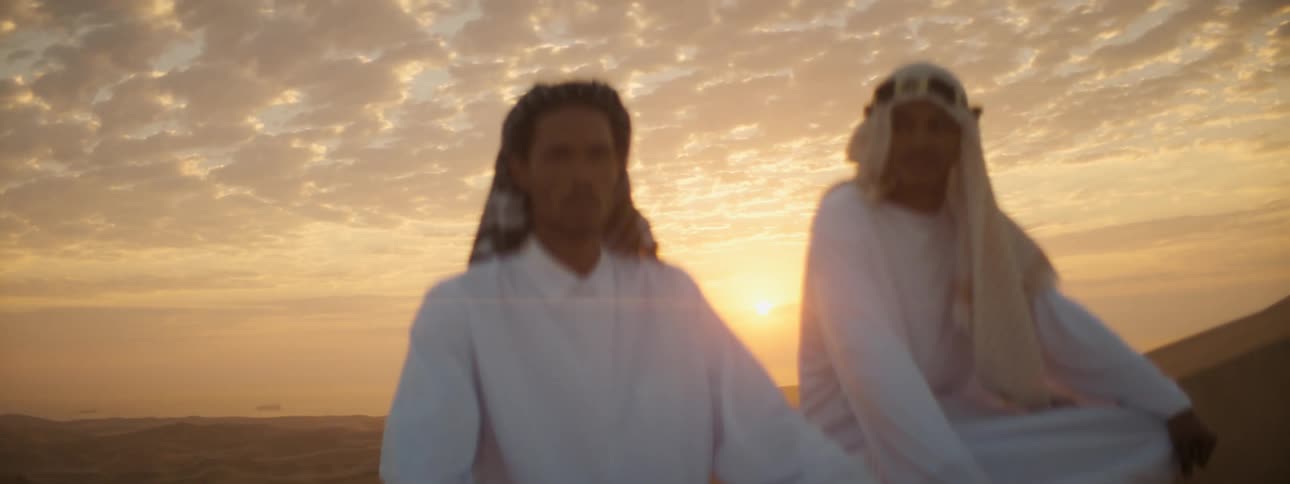 A pair of men in middle eastern clothing sit atop a sand dune in the desert in the evening