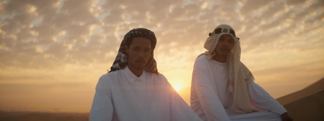 Two men sit in the sand at sunset