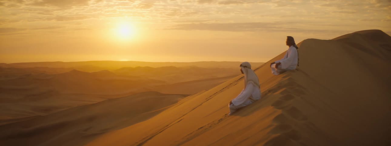 A pair of men sit atop a large dune in the desert at sunset and look out at the dunes below