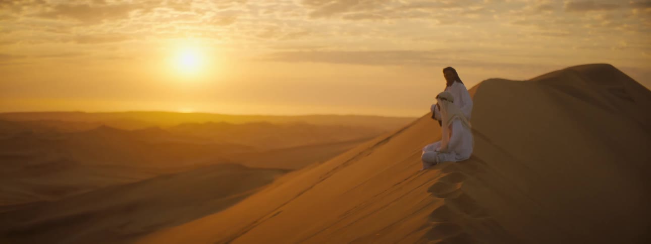 Two men sit on sand dune looking out onto desert landscape under sun in red sky