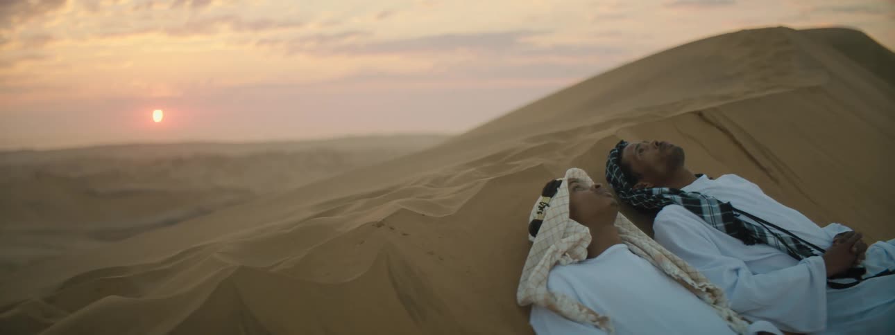 Two men laying on the desert sand with the sunset the sky and clouds in the background