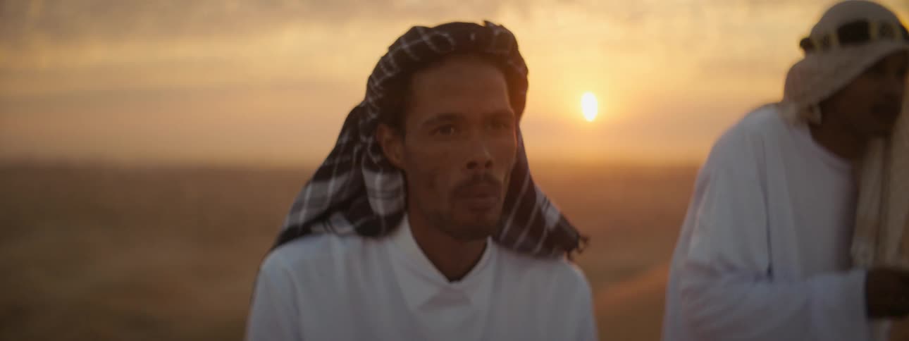 Two men sit on the sand in a desert and eat out of their hands as the sun sets