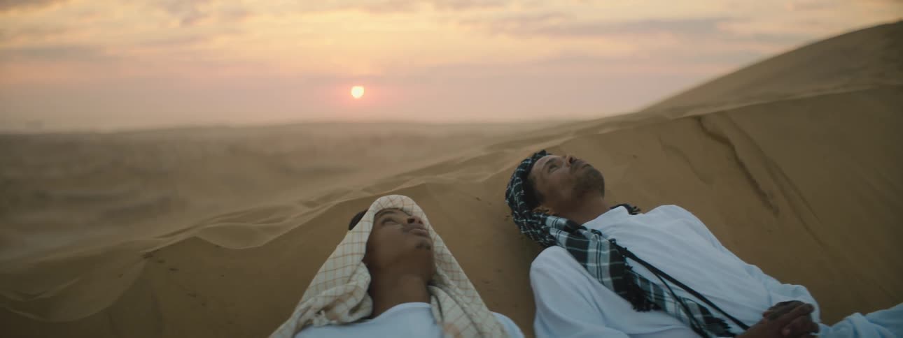 Two men lie on a sandy hill in the desert and look up at the sky
