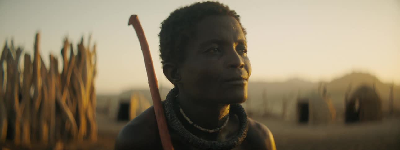 A black male standing in a hut village holding a stick