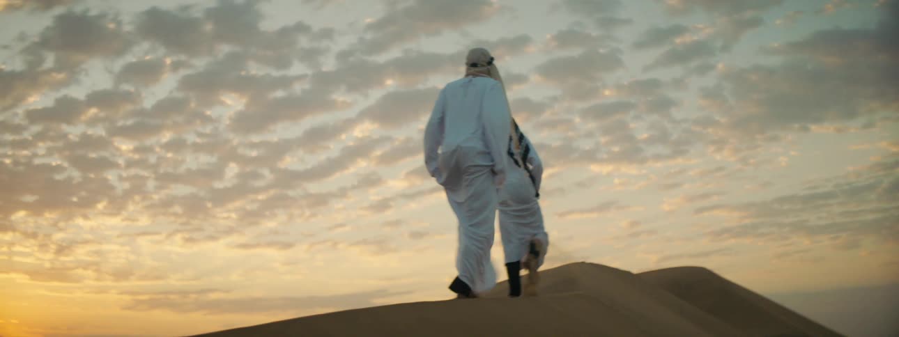 A pair of men in keffiyehs and thawbs walk across the top of a large sand dune in the desert at sunset