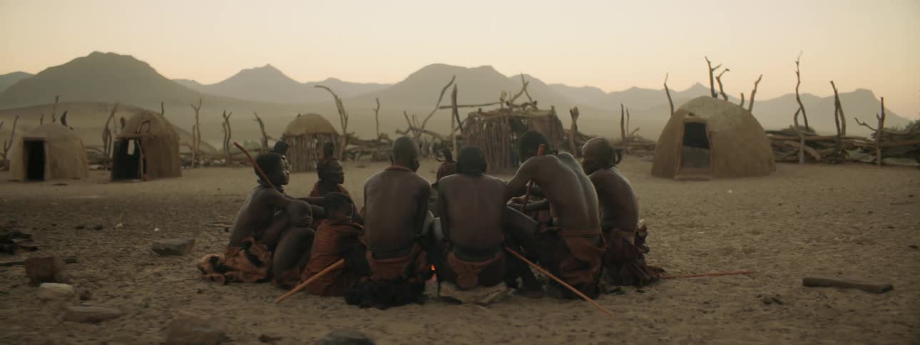 A group of tribal people sit in a circle around a fire during the day