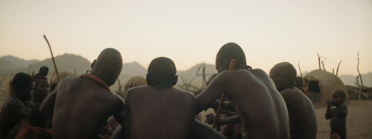 Villagers sits together in a group in fenced area containing huts with mountains on the horizon