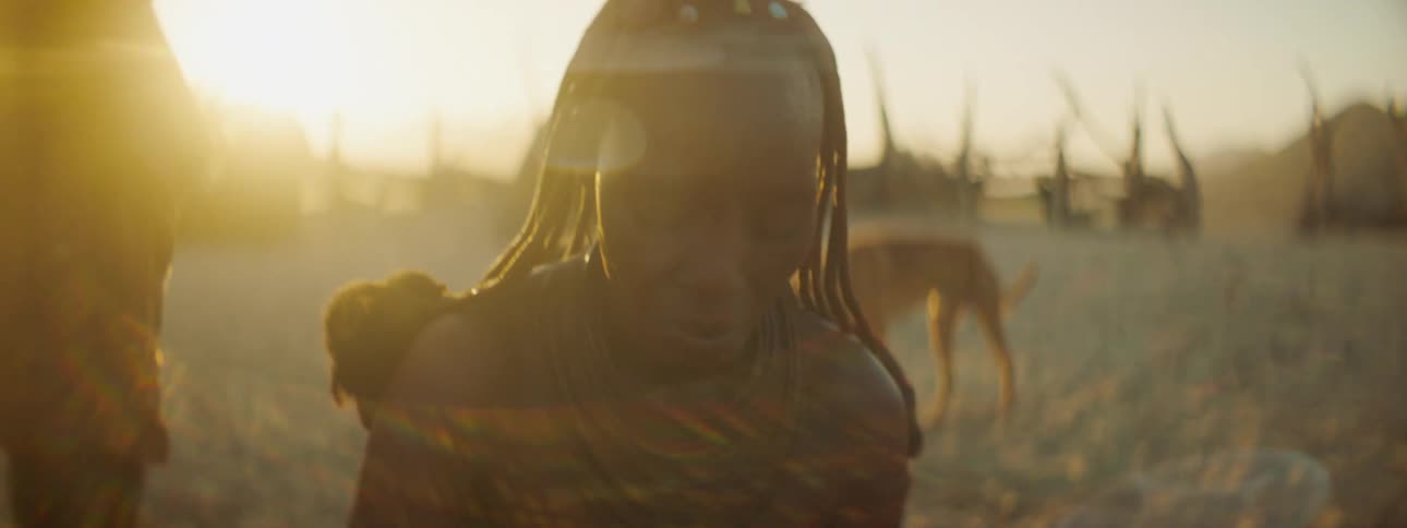 Women crouch on sandy ground back lit by setting sun as dog walks across sand in open area of village