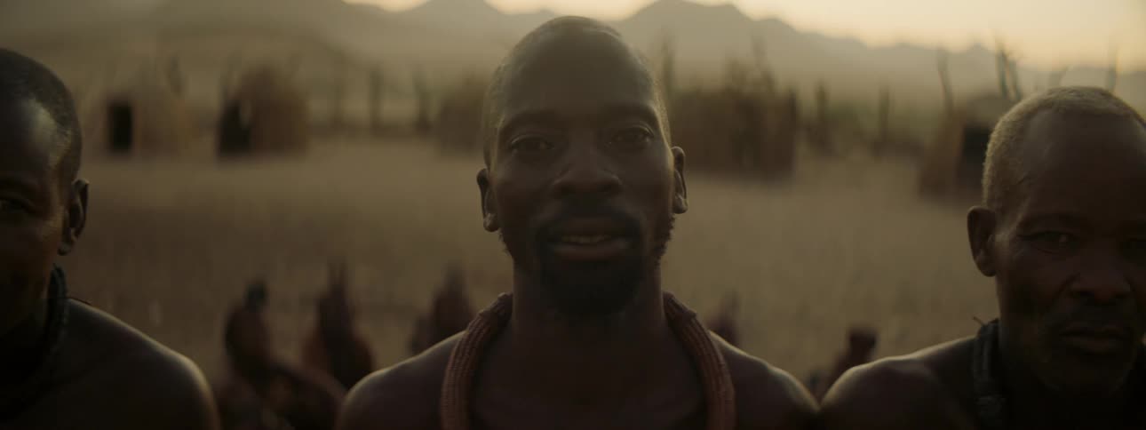 Men in a tribe stand in front of huts on a grassland