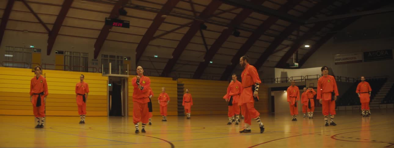 People in martial arts garb walk along court partly lit gym