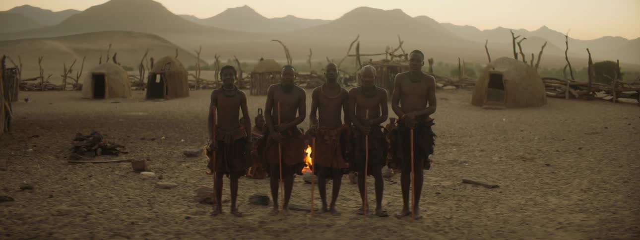 Tribe members stand in front of other members around a fire