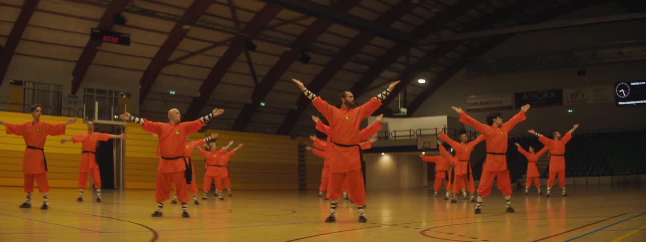 A group of people wearing red outfits are uniformly raising their arms over their heads in a gymnasium